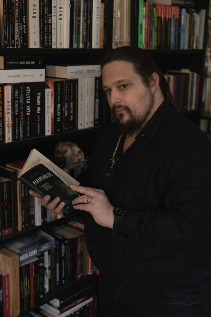 Michał Stonawski, portret amerykański, postać stojąca ubrana na czarno, zwrócona profilem, wzrok skierowany w kierunku obiektywu, w rękach przed sobą trzyma otwarty egzemplarz "Paranormalne. Prawdziwe historie nawiedzeń".