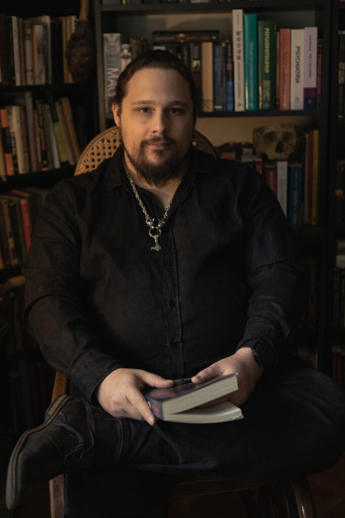 Michał Stonawski, portret amerykański, postać w czarnej koszuli w centrum, siedzi z założoną nogą na nogę i trzyma książkę w ręce. W tle biblioteczka.