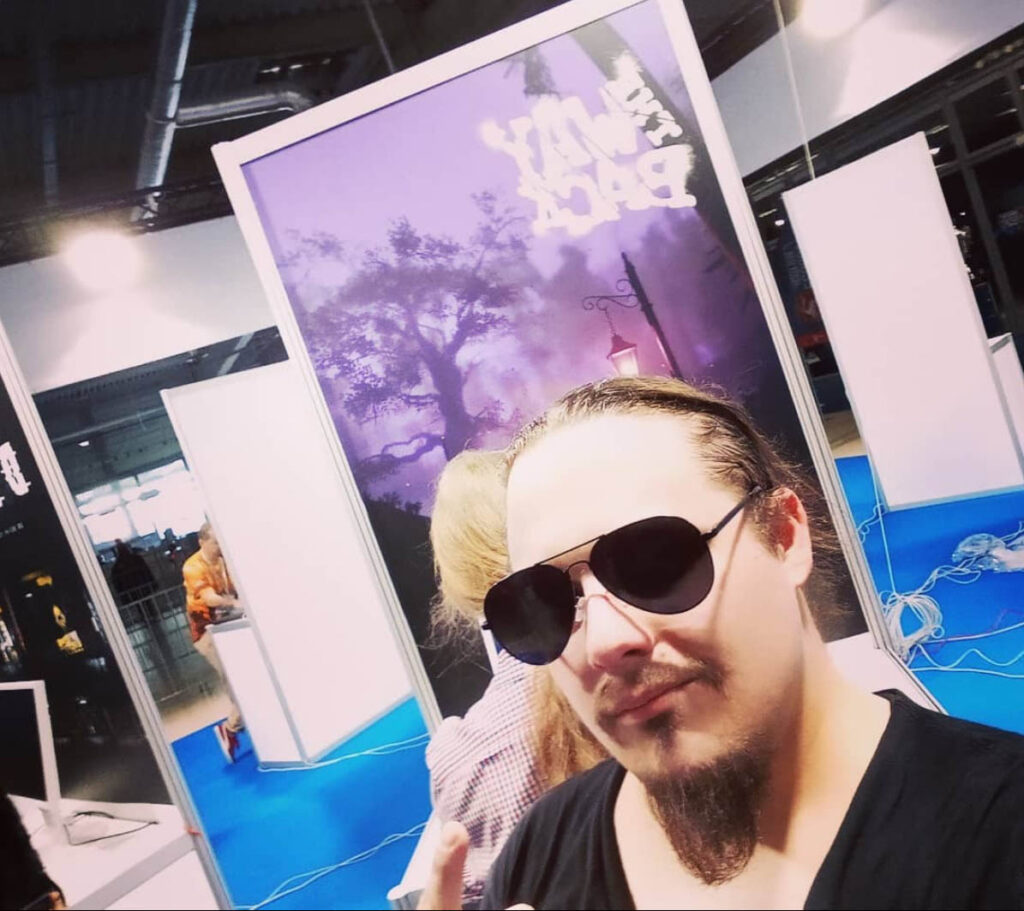 Na zdjęciu znajduje sie Michał Stonawski. Jest to zdjęcie typu selfie. Michał pozuje w czarnych dużych okularach na tle planszy z napisem The Way Back i screenem gry o tym tytule.